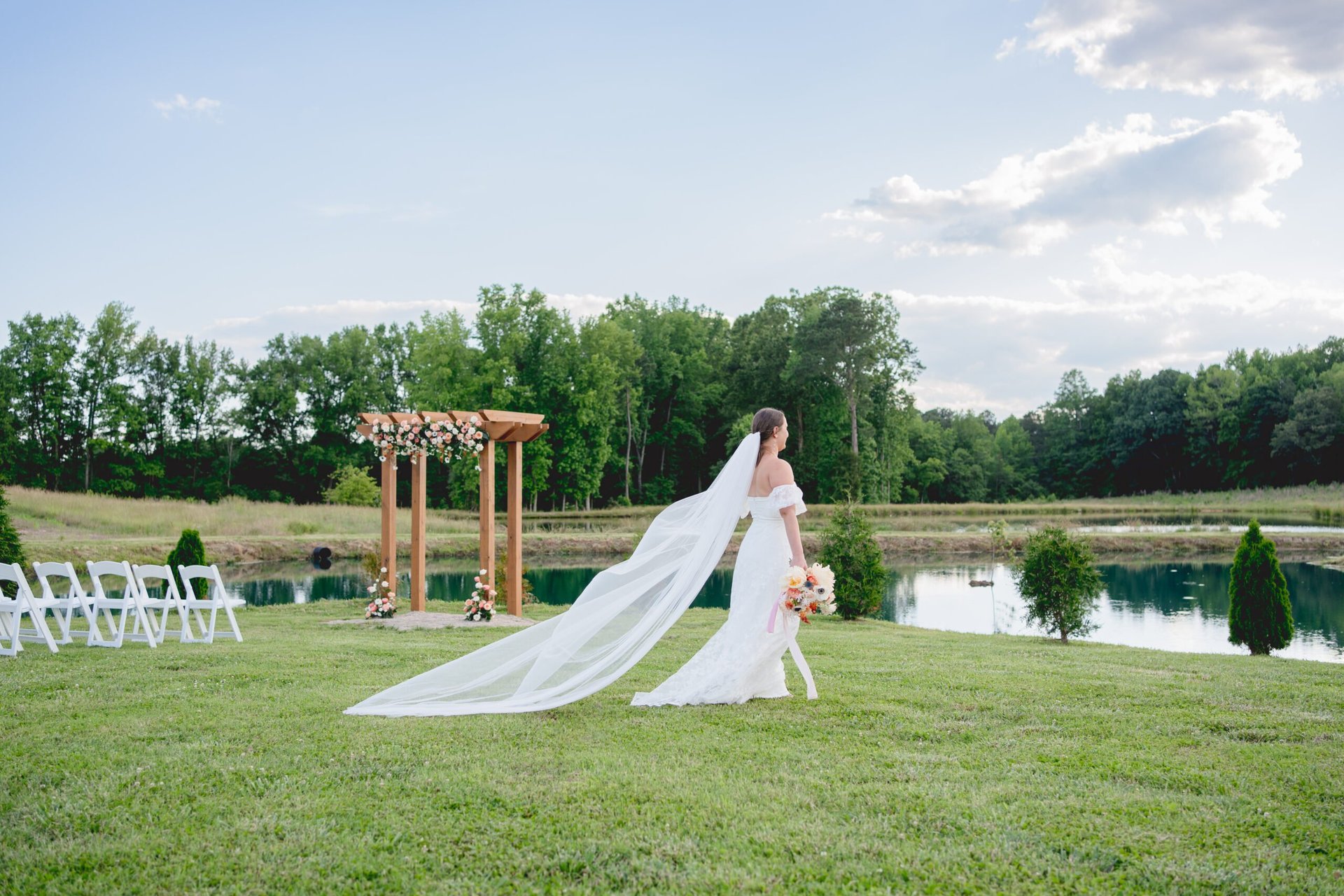 Bride in white wedding dress with long veil standing on grass near wooden arch by a pond